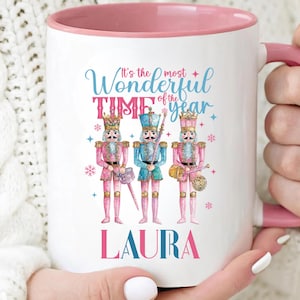 May include: White ceramic mug with a pink interior and handle. The mug features a festive design with three nutcracker soldiers and the text "It's the most Wonderful Time of the Year" and the name "LAURA".