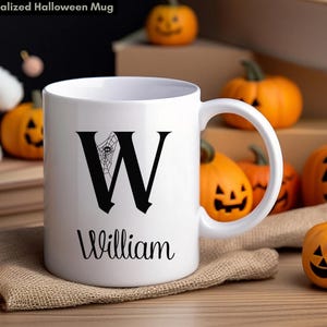 May include: White ceramic mug with a black letter "W" and a spiderweb design. The name "William" is printed below the letter. The mug is surrounded by small orange pumpkins with carved faces, perfect for Halloween.