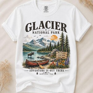 Può includere: Una t-shirt bianca a maniche corte con una grafica del Glacier National Park che mostra montagne, un lago, un alce e una canoa rossa. Il testo recita GLACIER NATIONAL PARK, WILD PLACES CLEAR SKIES GOOD TIMES e ADVENTURE IS OUT THERE MONTANA, USA.