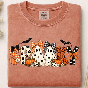 Könnte beinhalten: Ein terrakottafarbenes T-Shirt mit Rundhalsausschnitt und dem Wort SPOOKY in einer dekorativen Schriftart. Die Buchstaben sind mit orangefarbenem Vichy-Muster, schwarz-weißen Blumen, Leopardenmuster und Punkten gefüllt. Zwei Geister mit Schleifen und Kürbissen.