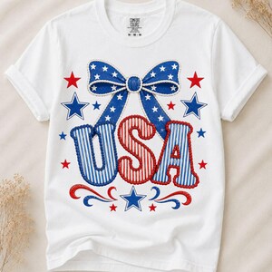 Könnte beinhalten: Ein weißes kurzärmeliges T-Shirt mit einem Grafikdruck einer blauen Schleife mit weißen Sternen über dem Text USA in roten und blauen Streifen. Das Design ist auf der Vorderseite des Shirts von verschiedenen roten und blauen Sternen umgeben.