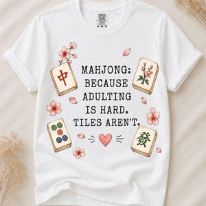 Könnte beinhalten: Ein weißes Kurzarm-T-Shirt mit dem schwarzen Text "MAHJONG: BECAUSE ADULTING IS HARD. TILES AREN'T.", umgeben von Illustrationen aus vier Mahjong-Steinen, rosa Kirschblüten und einem kleinen roten Herz, das unter dem Text zentriert ist.