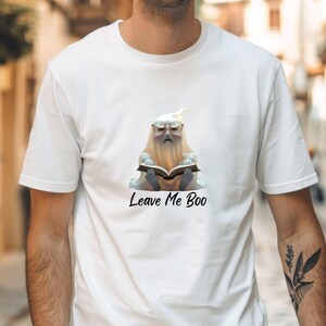 Puede incluir: Camiseta blanca con un fantasma de dibujos animados con gafas leyendo un libro. El fantasma tiene una expresión de mal humor. El texto "Leave Me Boo" está impreso debajo en cursiva negra.