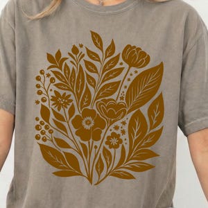 Könnte beinhalten: Graues T-Shirt mit einem braunen Blumendesign. Das Design zeigt verschiedene Blumen, Blätter und Beeren in kreisförmiger Anordnung. Das T-Shirt hat kurze Ärmel und einen Rundhalsausschnitt. Das Blumendesign ist braun.