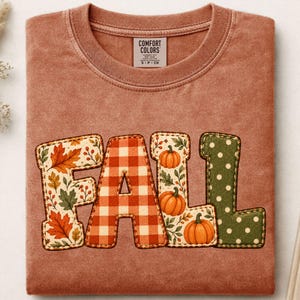 Könnte beinhalten: Ein terrakottafarbenes T-Shirt mit Rundhalsausschnitt und einem Comfort Colors-Etikett am Kragen. Auf der Vorderseite steht das Wort FALL in großen Blockbuchstaben, die jeweils mit verschiedenen Mustern bedruckt sind: Herbstblätter, orangefarbenes Vichy-Karo, Kürbisse und grüne Punkte.