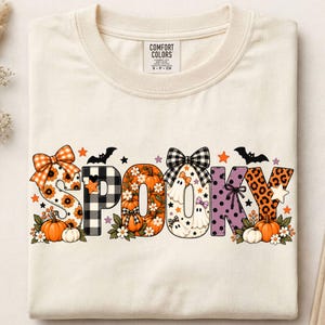 Könnte beinhalten: Ein cremefarbenes T-Shirt mit dem Wort SPOOKY in großen Buchstaben. Jeder Buchstabe zeigt einzigartige Muster, darunter orangefarbene Sonnenblumen, schwarz-weißes Karomuster, Kürbisse, Geister, violette Punkte und ein Leopardenmuster mit Schleifen.