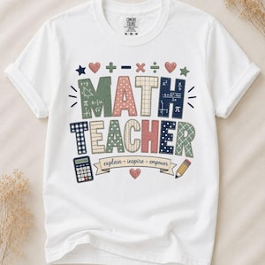 Könnte beinhalten: Ein weißes Kurzarm-T-Shirt mit dem Text MATH TEACHER in großen, gemusterten Buchstaben. Darunter befindet sich ein Banner mit der Aufschrift explain inspire empower, flankiert von einem Taschenrechner, einem Stift, Symbolen und Herzen.