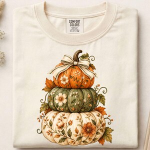Op de afbeelding: Een crèmekleurig t-shirt met een Comfort Colors-label in de nek. De afbeelding toont een stapel van drie pompoenen in oranje, groen en wit, versierd met bloemmotieven en ranken. Een gestreepte strik zit bovenop de pompoenen.