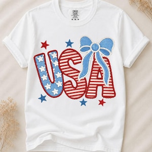 Könnte beinhalten: Ein weißes kurzärmeliges T-Shirt mit einer großen Grafik der Buchstaben USA. Das U ist blau mit weißen Sternen, das S hat rote und weiße horizontale Streifen und das A ist mit einer großen hellblauen Schleife verziert.