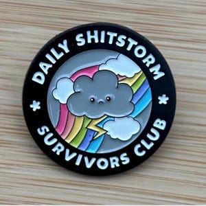 May include: A round enamel pin with a black border featuring the white text DAILY SHITSTORM SURVIVORS CLUB. The centre depicts a sad grey cloud with a face, white clouds, a colourful rainbow, and a yellow lightning bolt on a light grey background.
