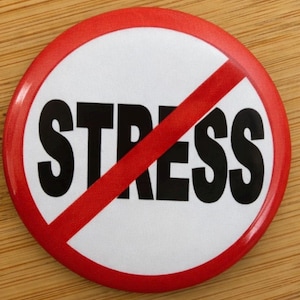 May include: A circular badge with a white centre and a red border. The word "STRESS" is printed in bold black letters, with a red diagonal line through it. The badge is on a wooden surface.