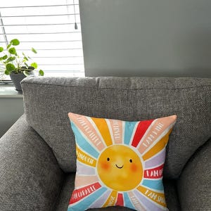May include: Square cushion with a smiling sun design and colourful rays. The rays feature positive affirmations like "I am kind" and "I am enough". The cushion is placed on a grey upholstered armchair.