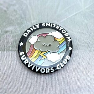 May include: A circular enamel pin with the words "DAILY SHITSTORM SURVIVORS CLUB" in white on a black border. The pin shows a cartoon cloud, rainbow, and lightning.