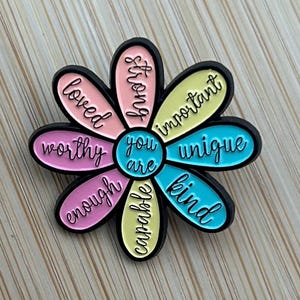 May include: A flower-shaped enamel pin with black edging. The petals are in shades of pink, yellow, and blue, each with a positive word such as "loved", "strong", and "unique". The centre of the flower reads "you are".