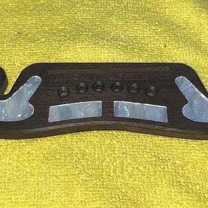 May include: A dark brown guitar bridge with decorative mother-of-pearl inlays. The bridge has a curved shape with six holes for strings and two decorative swirls on each end. The inlays are rectangular and curved.