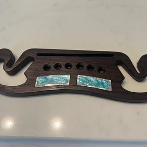 May include: A dark brown wooden guitar bridge featuring a decorative inlay of two rectangular turquoise-colored pieces with iridescent borders. The bridge has a long horizontal slot and five circular holes arranged in a row across the center.