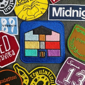 Op de afbeelding: Een verzameling kleurrijke geborduurde patches met verschillende ontwerpen, waaronder een huis met een regenboogkleurenschema, een hartvorm, nummer 89, een stopbord met de tekst "Taylor's Version" en het woord "Midnight".