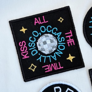 May include: Black square patch with embroidered text and a disco ball design. The text reads "Kiss Disco Occasionally All The Time" in pink, blue, and yellow. Includes yellow star accents.