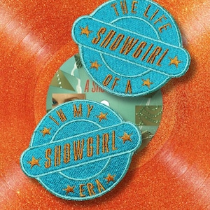 Könnte beinhalten: Zwei gestickte Aufnäher mit dem Text "SHOWGIRL" in Orange, umgeben von einem blauen Kreis und Sternen. Ein Aufnäher lautet "THE LIFE OF A SHOWGIRL" und der andere "IN MY SHOWGIRL ERA". Die Aufnäher befinden sich auf einem orangefarbenen Glitzerhintergrund.