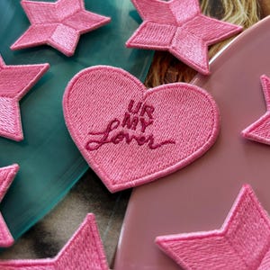 Puede incluir: Parche bordado en forma de corazón rosa con el texto "UR MY LOVER" en hilo rosa. El parche está rodeado de parches bordados en forma de estrella rosa.
