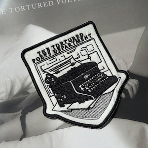 Pode incluir: Etiqueta branca bordada com pontos pretos que representa uma máquina de escrever e o texto "The Tortured Poets Department".