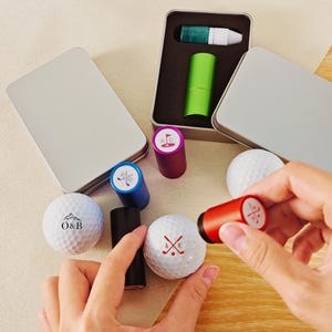 Puede incluir: Juego de sellos para pelotas de golf con iniciales personalizadas y diseños temáticos de golf. Incluye un sello rojo, azul y negro, pelotas de golf y un rotulador verde, todo en una lata plateada. Los sellos presentan iniciales y gráficos de palos de golf.