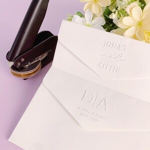 May include: A black and silver seal press beside two white envelopes with embossed text. One envelope reads "Jonas and Kittie", the other reads "D/A Donald & Arvin May 3 2025". Flowers are in the background.