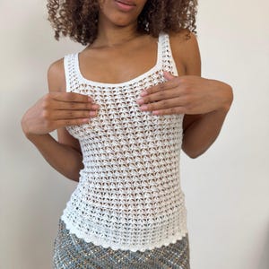 May include: A white, open-weave crochet tank top with a scoop neckline and a scalloped hem. The top is sleeveless and appears to be handmade. The crochet pattern creates a delicate, airy texture, perfect for warm weather.