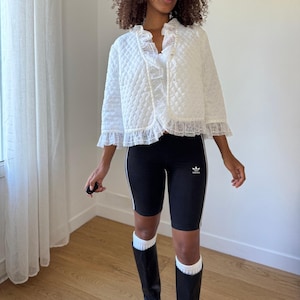 Könnte beinhalten: Eine weiße Steppjacke mit Spitzenbesatz, kombiniert mit schwarzen Sporthosen mit weißen Streifen und einem kleinen Logo. Das Outfit wird durch schwarze Stiefel und weiße Socken ergänzt.
