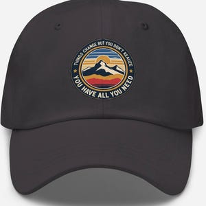May include: A dark gray baseball cap with a circular graphic featuring a mountain range, sun, and text that reads "THINGS CHANGE BUT YOU DON'T REALIZE YOU HAVE ALL YOU NEED". The design includes shades of blue, orange, and yellow.
