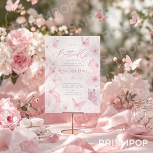 An elegant image featuring a delicate baby shower invitation elegantly displayed on a gold stand amid soft pink silk fabric, blush-toned roses, white blossoms, greenery, and gentle pastel butterflies gracefully floating in the background. The invitation showcases refined pink watercolor butterflies and florals, with sophisticated typography reading, "A Little Butterfly is on the Way," celebrating Wanessa Tang. Soft natural lighting and blurred botanical details lend a serene, airy.