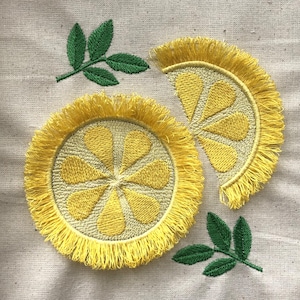 Pode incluir: Duas fatias de limão bordadas de amarelo com bordas franjadas, uma inteira e uma metade, com folhas bordadas de verde sobre um fundo de tecido bege.