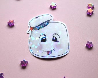 May include: A white iridescent sticker of the Stay Puft Marshmallow Man from Ghostbusters. The sticker has a holographic finish and features the character's face with a mischievous grin.