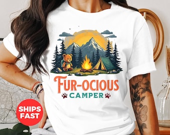 Puede incluir: Camiseta blanca con un diseño de camping. El diseño presenta un oso de peluche, una fogata, una tienda de campaña y montañas bajo un atardecer. El texto "FUR-OCIOUS CAMPER" está en naranja.