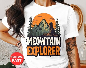May include: White t-shirt featuring a graphic of a cat sitting on a rock, with a mountain and sunset backdrop. The text "MEOWTAIN EXPLORER" is printed below the image in white and orange block letters. The design evokes a nature and adventure theme.