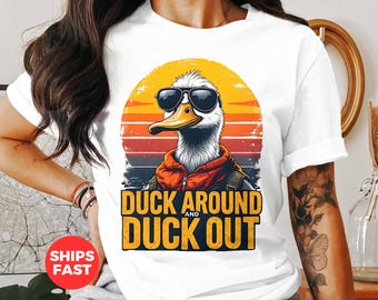 May include: White t-shirt featuring a graphic of a duck wearing sunglasses and a red jacket. The duck is in front of a sunset with the text "Duck Around and Duck Out".