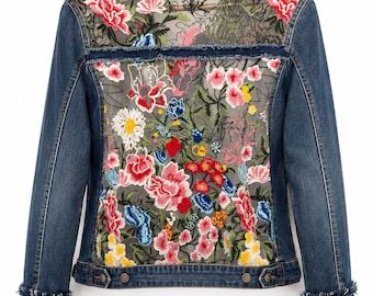 May include: A blue denim jacket featuring a sheer back panel with vibrant floral embroidery in shades of pink, red, yellow, and blue. The collar and cuffs are finished with frayed edges and a decorative trim of white faux pearls and dark thread.