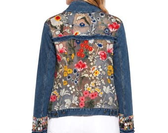 May include: A blue denim jacket featuring a sheer back panel with a vibrant floral print in red, yellow, and white. The jacket includes long sleeves with frayed cuffs decorated with small reflective mirror accents and colorful embroidery.