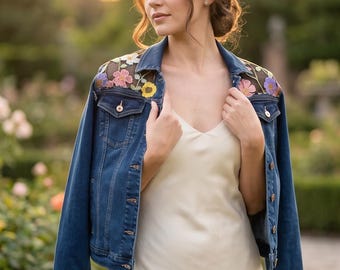 May include: A dark blue denim jacket featuring sheer black mesh panels on the shoulders adorned with colorful floral embroidery. The cuffs are decorated with matching floral embroidery and small silver studs. The jacket has a classic collar and buttons.