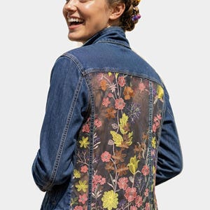 May include: A blue denim jacket featuring a large, sheer mesh back panel adorned with intricate, multi-colored floral embroidery in shades of pink, yellow, orange, and brown. The jacket has a classic collar, long sleeves, and metal button closures.