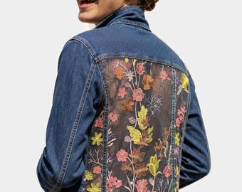 May include: A blue denim jacket featuring a large, sheer mesh back panel adorned with intricate, multi-colored floral embroidery in shades of pink, yellow, orange, and brown. The jacket has a classic collar, long sleeves, and metal button closures.