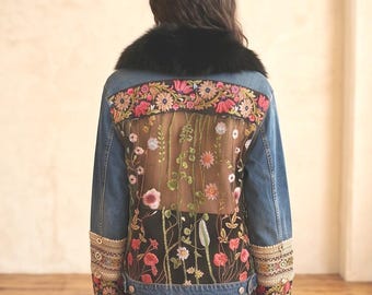 May include: A blue denim jacket featuring a black faux fur collar, long sleeves with intricate embroidered cuffs, and a back panel composed of sheer mesh and solid black fabric adorned with colorful floral embroidery and green vine detailing.