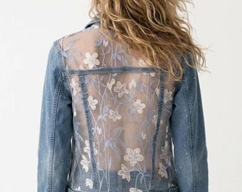 May include: A blue denim jacket featuring a sheer mesh back panel with light blue and white floral embroidery. The jacket includes long sleeves, a classic collar, metal button closures, and a structured denim hemline that sits at the waist.
