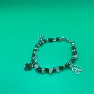 Könnte beinhalten: Ein Armband mit grünen und klaren Perlen und zwei silberfarbenen vierblättrigen Kleeblatt-Anhängern. Das Armband befindet sich auf einem grünen Hintergrund.