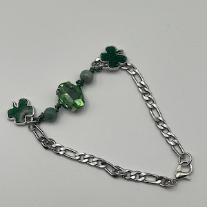 May include: A silver-toned bracelet featuring a chain link design. The bracelet is adorned with green shamrock charms and green beads. A large, faceted green crystal is also incorporated into the design. The bracelet has a lobster clasp closure.