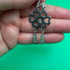 Op de afbeelding: Zilverkleurige oorhangers met een klaverontwerp met vier bladeren. Elke oorbel heeft een klavervormige bovenkant en twee kleine, ronde, groene kralen die eronder hangen. De oorbellen worden getoond tegen een felgroene achtergrond.