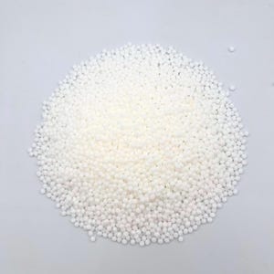 Puede incluir: Un montón de pequeñas cuentas blancas y esféricas. Las cuentas están densamente empaquetadas, formando una forma redondeada contra un fondo liso y de color claro. La textura parece suave y uniforme.