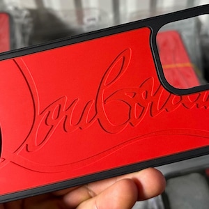 May include: Red phone case with a black border and the words "christian louboutin paris" printed on the bottom left corner. The case has a raised, embossed design of the words "louboutin" in a white font.
