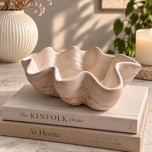 May include: A decorative bowl shaped like a clam shell with a smooth, off-white and light brown marbled finish. The bowl features a wavy, organic rim and rests on top of two stacked books titled The KINFOLK Home and At Home A GUIDE TO SIMPLE LIVING.