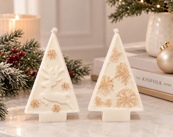 May include: Two triangular white candles shaped like Christmas trees. One candle features a raised white reindeer figure and small gold snowflake accents. The second candle is decorated with multiple gold snowflake embellishments. Both candles have a single wick at the top.
